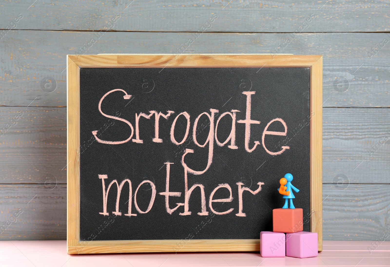 Photo of MYKOLAIV, UKRAINE - JANUARY 04, 2022: Small blackboard with words Surrogate Mother, cubes and human figures on pink wooden table