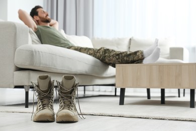 Photo of Soldier relaxing on sofa in living room, focus on pair of combat boots. Military service