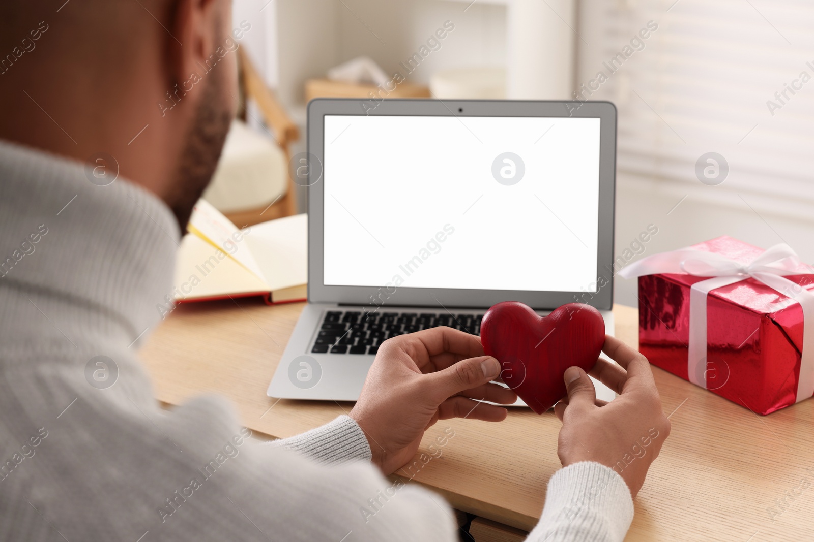 Photo of Valentine's day celebration in long distance relationship. Man holding red wooden heart while having video chat with his girlfriend via laptop, closeup