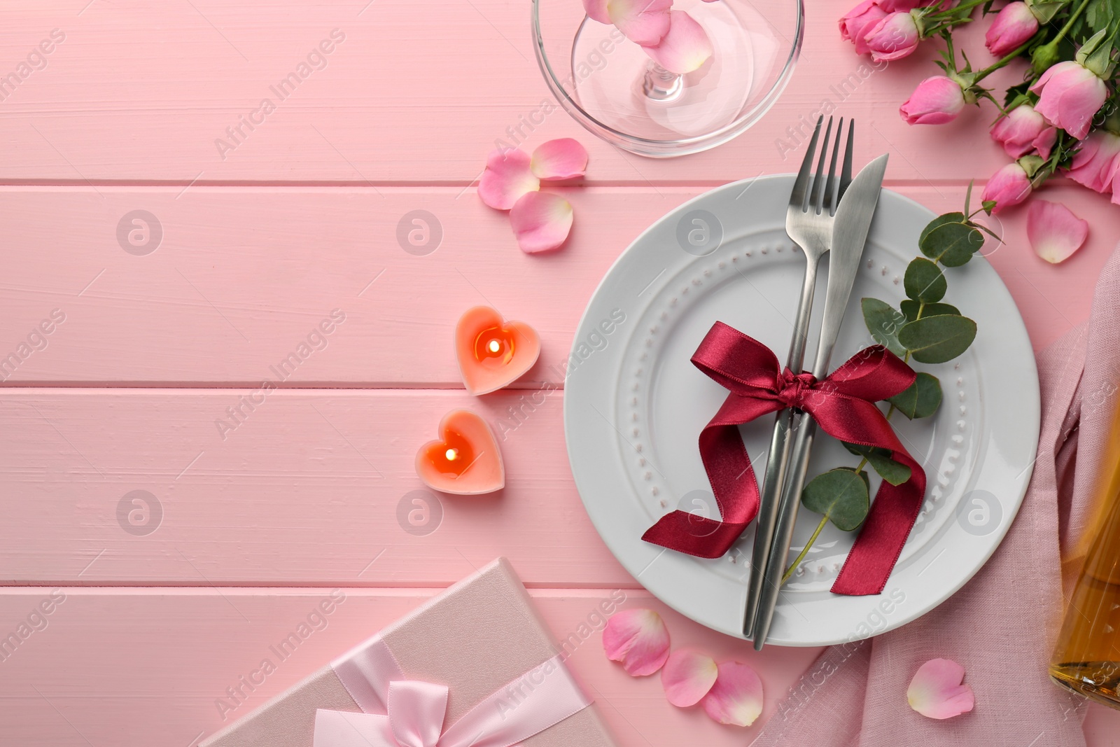 Photo of Place setting with heart shaped candles, bouquet of roses and gift for romantic dinner on pink wooden table, flat lay. Space for text