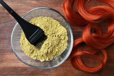 Photo of Bowl of henna powder, brush and red strand on wooden table, flat lay. Natural hair coloring