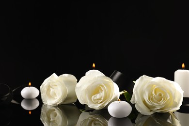 Photo of White roses and burning candles on black mirror surface in darkness, space for text. Funeral symbols