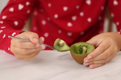 Photo of Girl eating tasty fresh kiwi with spoon at white marble table indoors, closeup