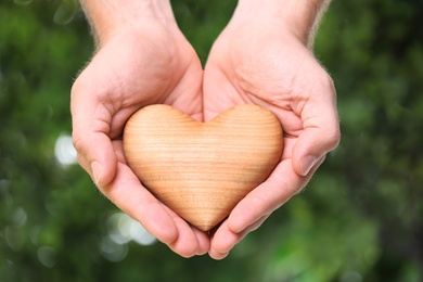 Young man holding wooden heart on blurred green background, closeup. Donation concept