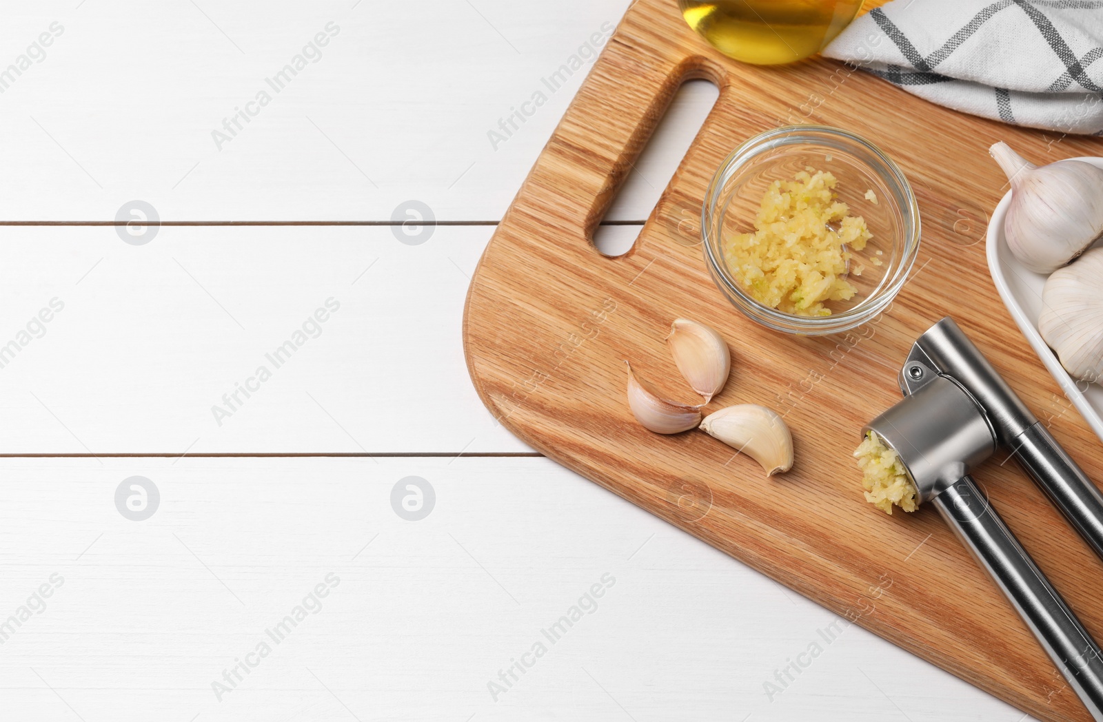 Photo of Board with metal press and garlic on white wooden table, top view. Space for text