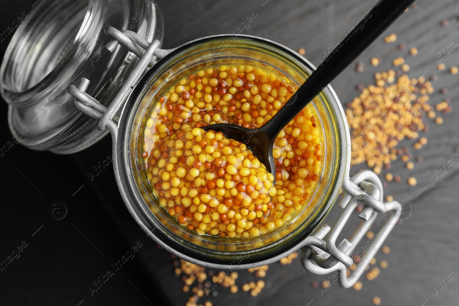 Photo of Whole grain mustard and spoon in jar on black wooden table, top view