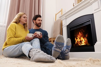 Lovely couple with hot drinks spending time together near fireplace at home, low angle view