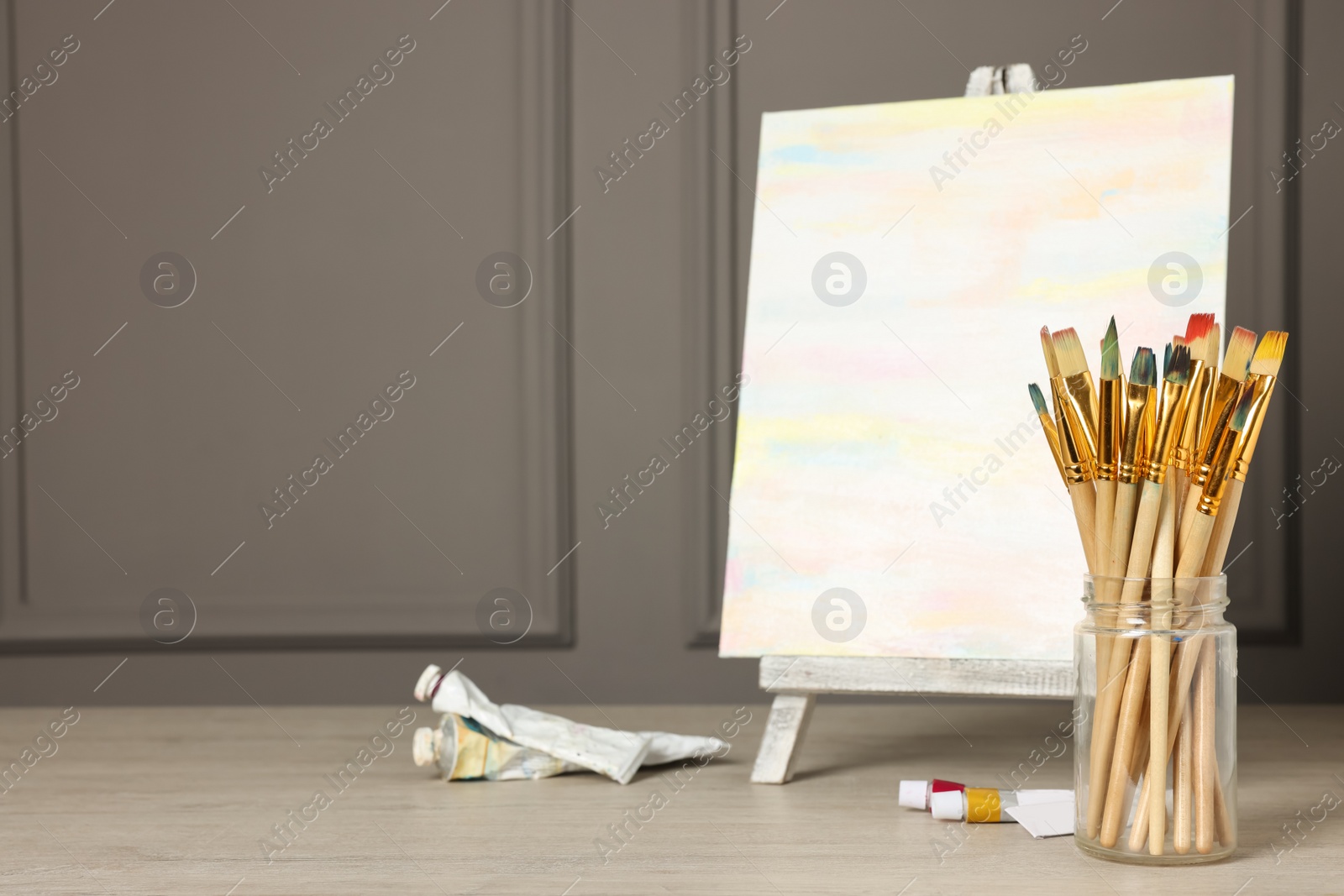 Photo of Different brushes, easel with abstract picture and paints on wooden table. Space for text