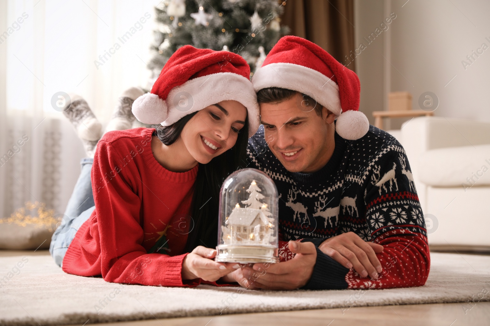 Photo of Couple in Santa hats holding snow globe at home