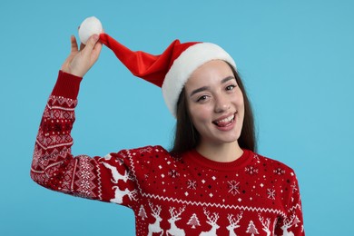 Photo of Happy young woman in Christmas sweater and Santa hat on light blue background