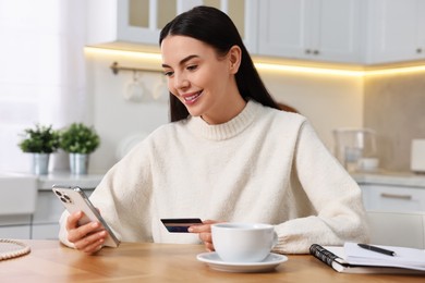 Happy young woman with smartphone and credit card shopping online at wooden table in kitchen