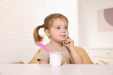 Cute little child eating tasty yogurt from plastic cup with spoon at white table indoors