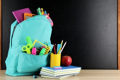 Bright backpack with school stationery on brown wooden table near black chalkboard, space for text