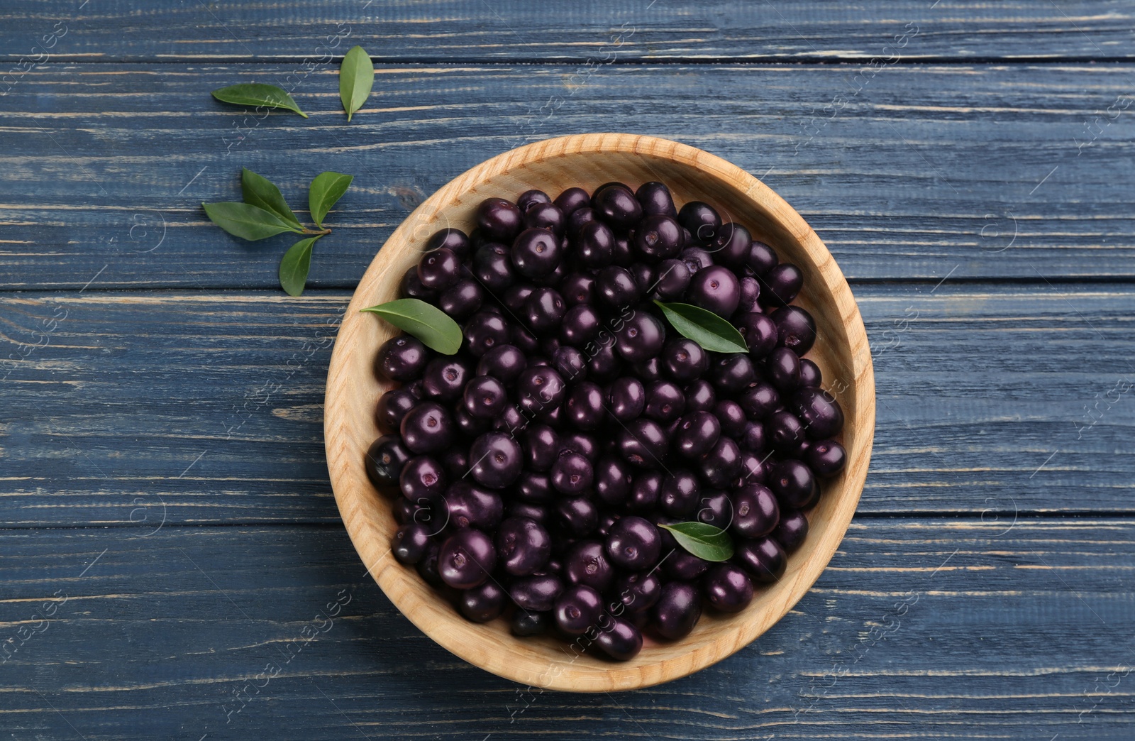 Photo of Fresh acai berries in bowl and leaves on blue wooden table, flat lay