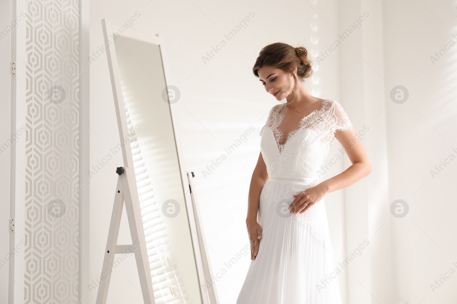 Photo of Young bride in beautiful wedding dress near mirror indoors