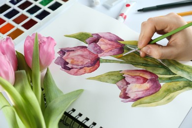 Photo of Woman painting tulips in sketchbook at white table, closeup