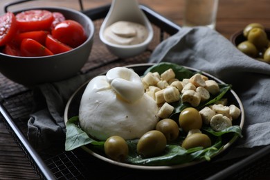 Photo of Delicious burrata cheese served with olives, croutons, basil and tomatoes on wooden table, closeup