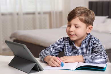 Little boy erasing mistake in his notebook near tablet at white desk indoors