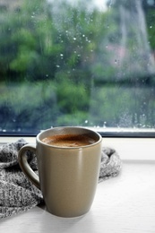 Photo of Cup of hot drink on window sill against glass with rain drops, space for text