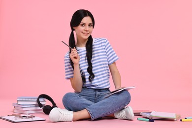 Smiling student with notebook sitting among books and stationery on pink background