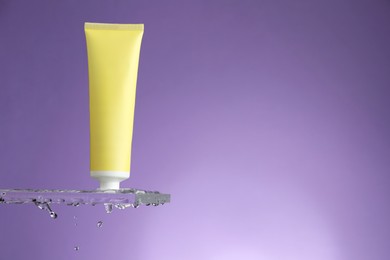 Photo of Moisturizing cream in tube on glass with water drops against violet background. Space for text