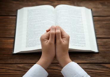 Photo of Woman holding hands clasped while praying at wooden table with Bible, closeup