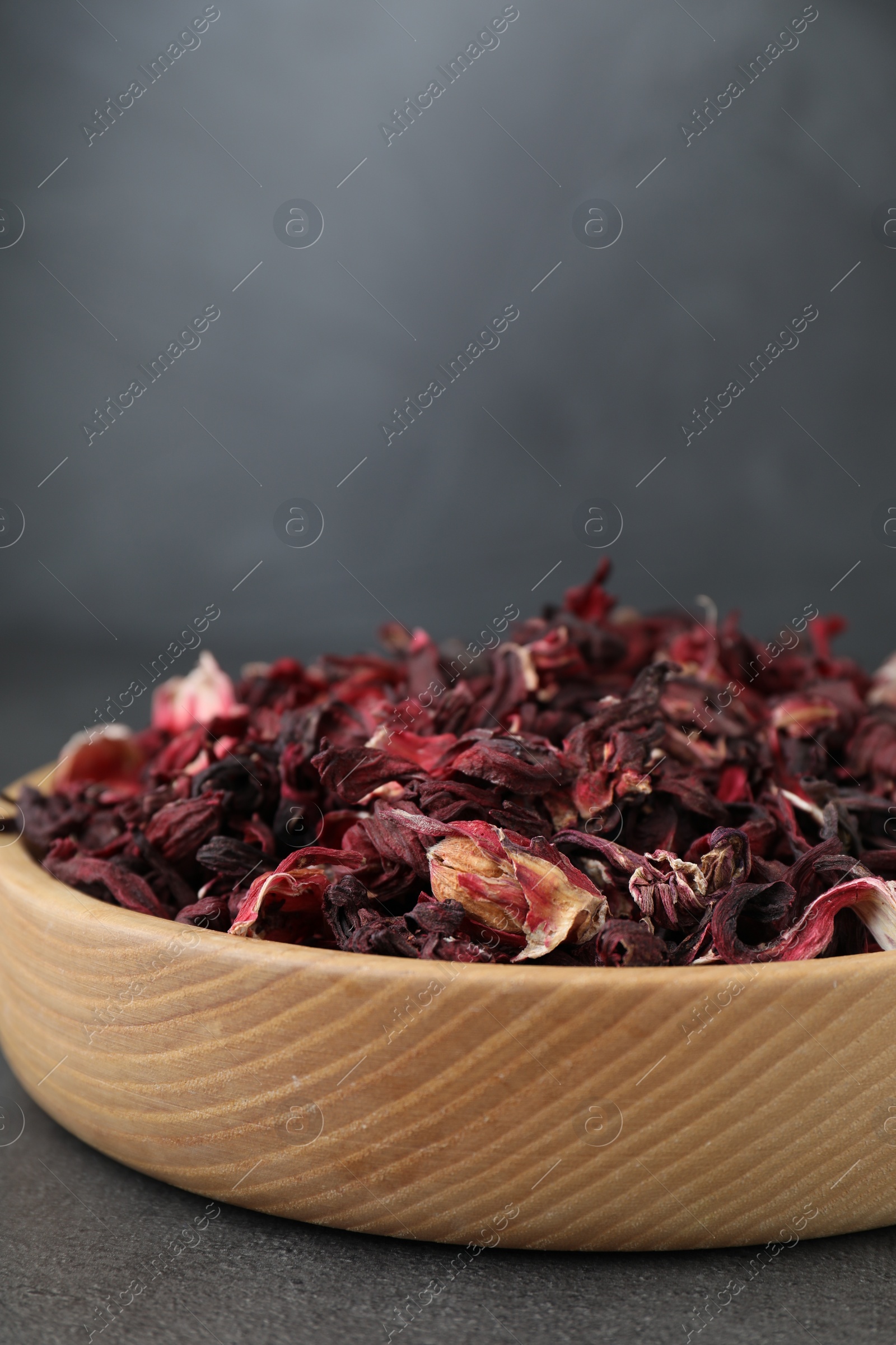 Photo of Hibiscus tea. Wooden bowl with dried roselle calyces on grey table, closeup. Space for text