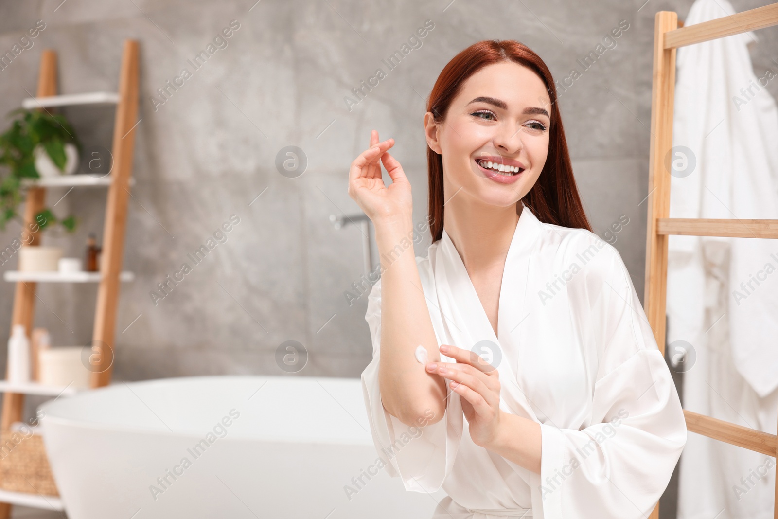 Photo of Beautiful young woman applying body cream onto elbow in bathroom, space for text