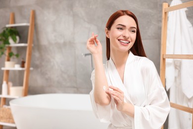 Beautiful young woman applying body cream onto elbow in bathroom, space for text