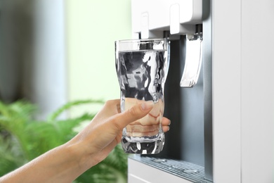 Photo of Woman filling glass from water cooler indoors, closeup. Refreshing drink