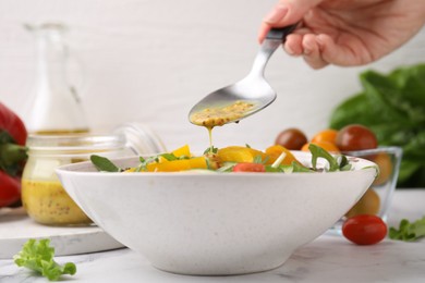 Photo of Woman pouring tasty vinegar based sauce (Vinaigrette) from spoon into bowl with salad at white marble table, closeup