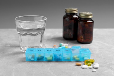 Photo of Weekly pill box with medicaments and glass of water on grey marble table