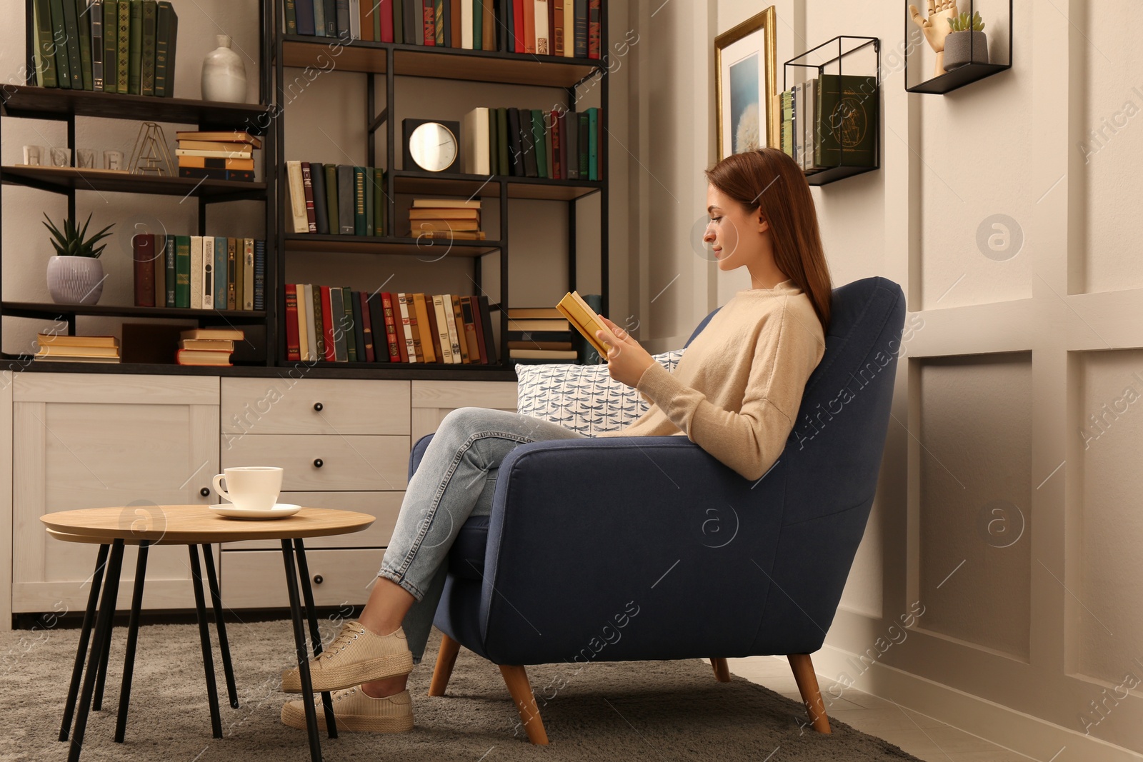 Photo of Young woman reading book in armchair indoors. Home library