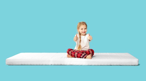 Photo of Little girl sitting on mattress and showing thumbs up against light blue background