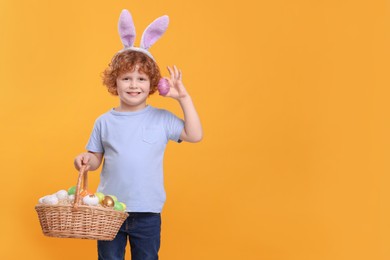 Photo of Portrait of happy boy in cute bunny ears headband holding wicker basket with Easter eggs on orange background. Space for text