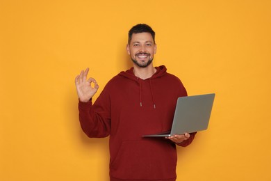 Smiling man with laptop showing okay gesture on orange background