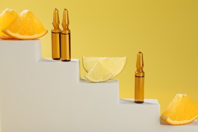 Stylish presentation of skincare ampoules with vitamin C and citrus slices on decorative stairs against yellow background, closeup