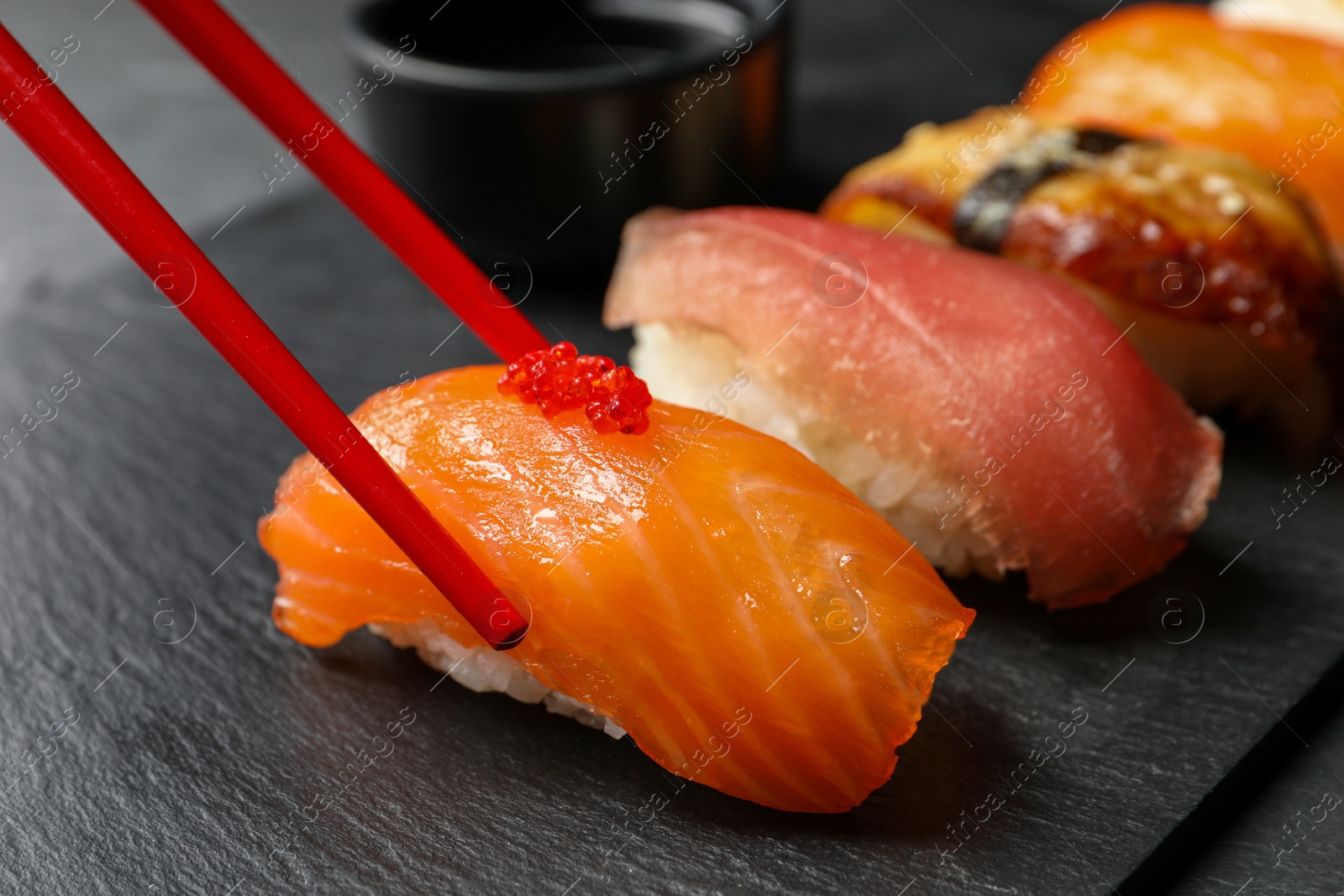 Photo of Taking delicious nigiri sushi with chopsticks from serving board on black table, closeup