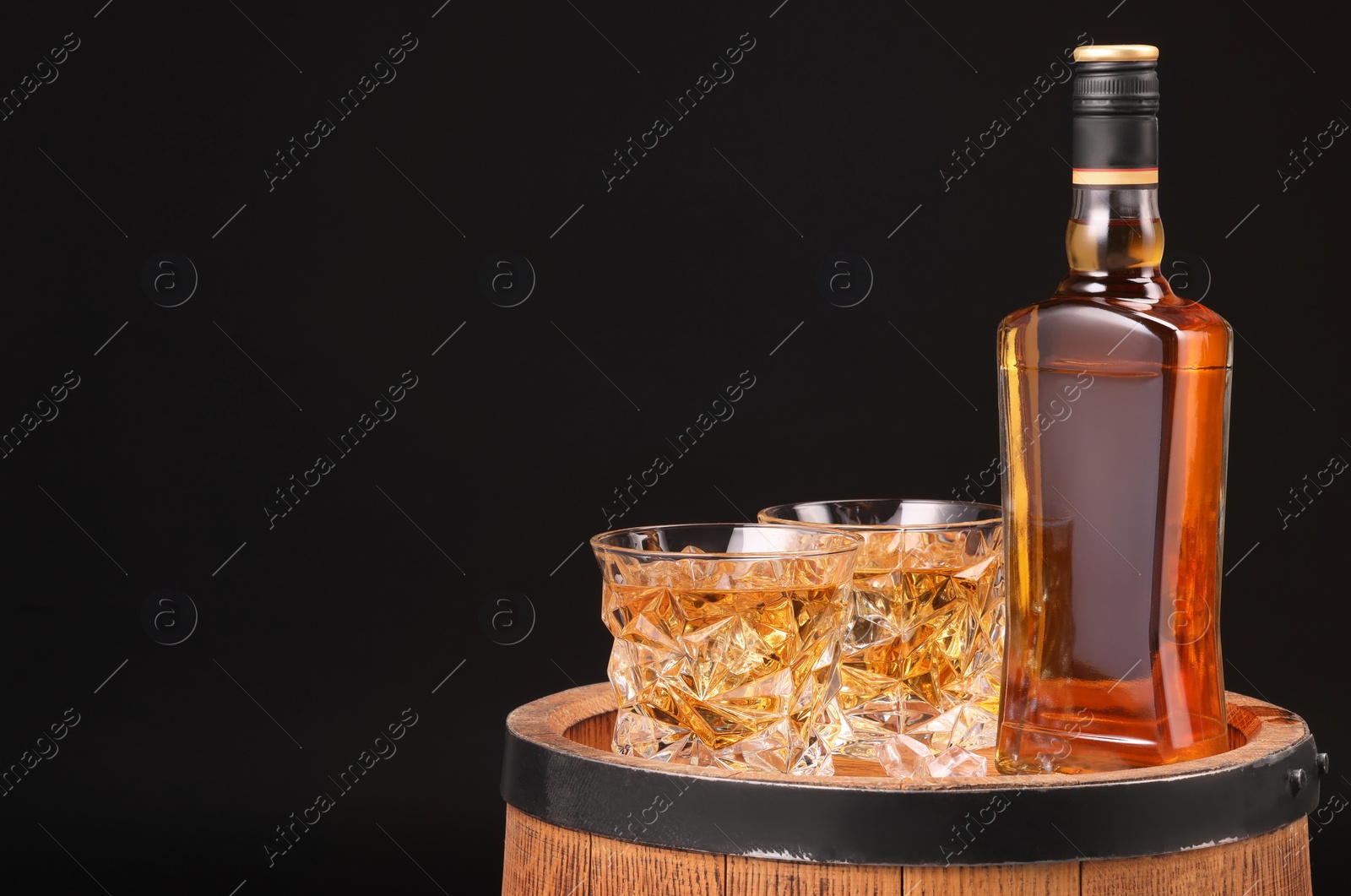 Photo of Whiskey in glasses and bottle on wooden barrel against black background, space for text