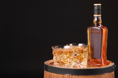 Whiskey in glasses and bottle on wooden barrel against black background, space for text