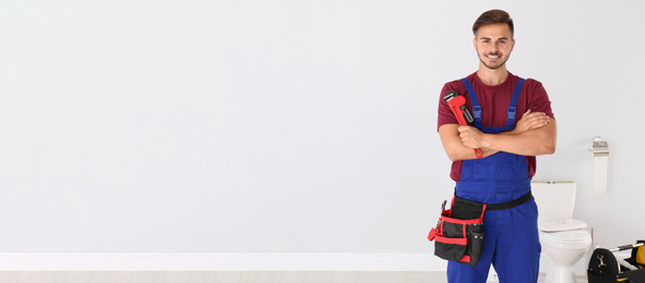 Young man with plumber wrench near toilet bowl indoors, space for text. Banner design