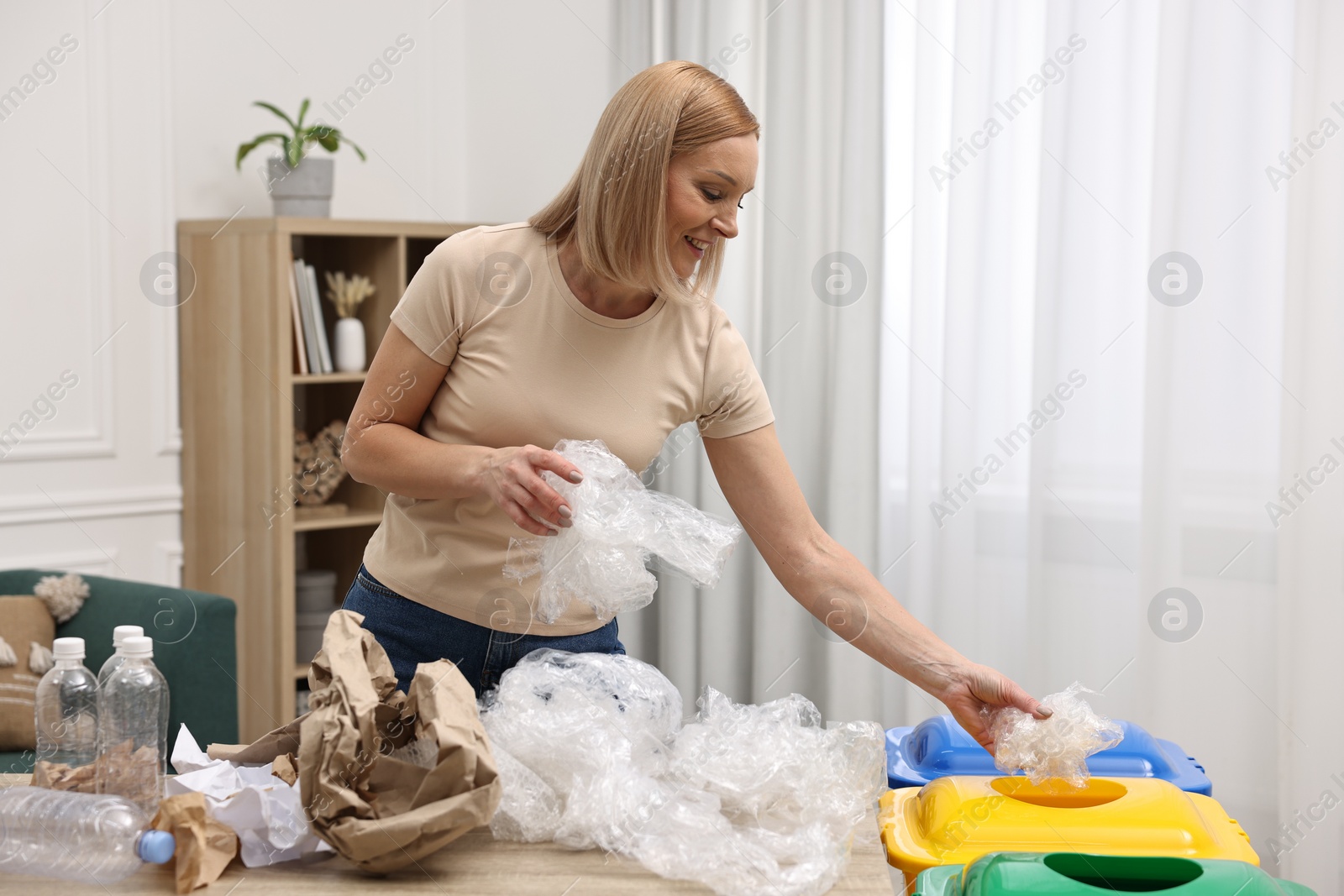 Photo of Garbage sorting. Smiling woman throwing plastic package into trash bin in room