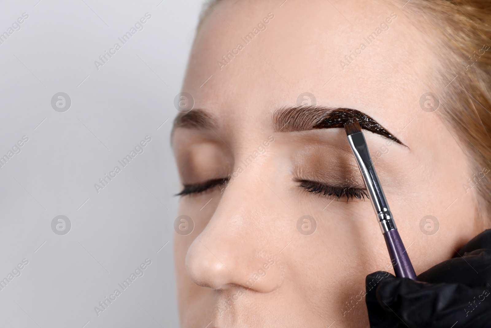 Photo of Beautician applying tint during eyebrows correction procedure on light grey background, closeup