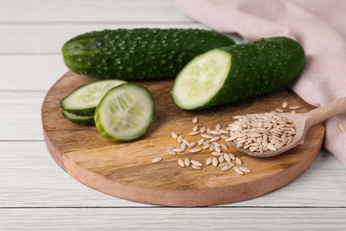 Photo of Spoon of vegetable seeds and fresh cucumbers on white wooden table, closeup