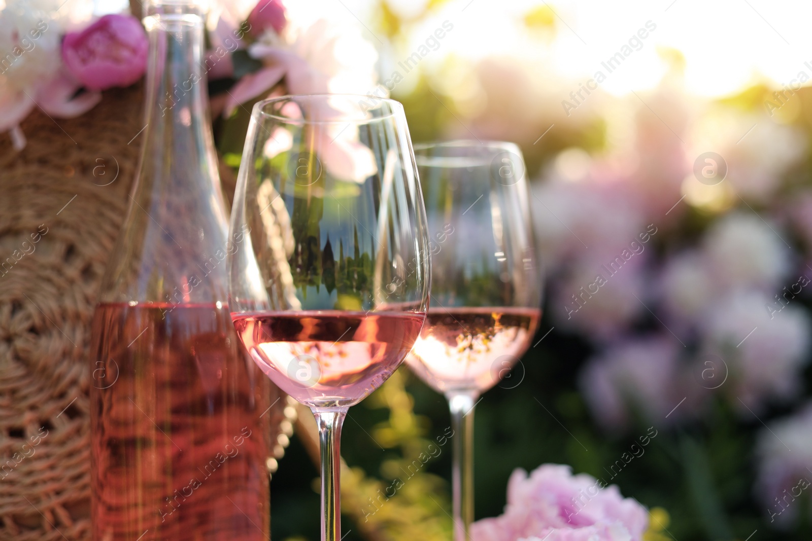 Photo of Bottle and glasses of rose wine near straw bag with beautiful peonies in garden, closeup