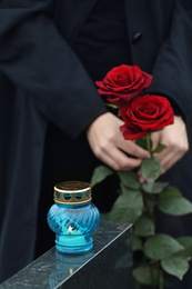 Photo of Woman holding red roses near black granite tombstone with candle outdoors, closeup. Funeral ceremony