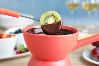 Photo of Dipping kiwi slice into pot with chocolate fondue on table, closeup