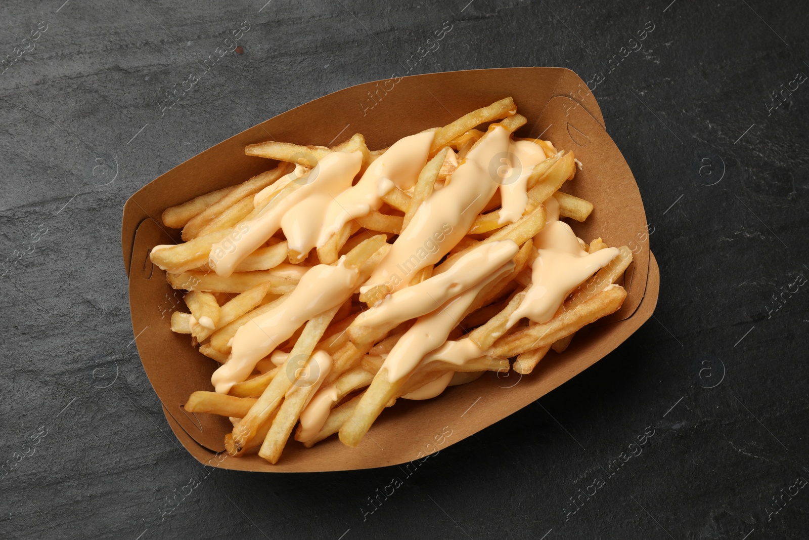 Photo of Tasty potato fries and cheese sauce in paper container on black table, top view