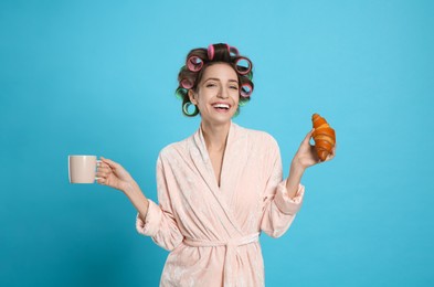 Photo of Happy young woman in bathrobe with hair curlers holding croissant and cup of drink on light blue background