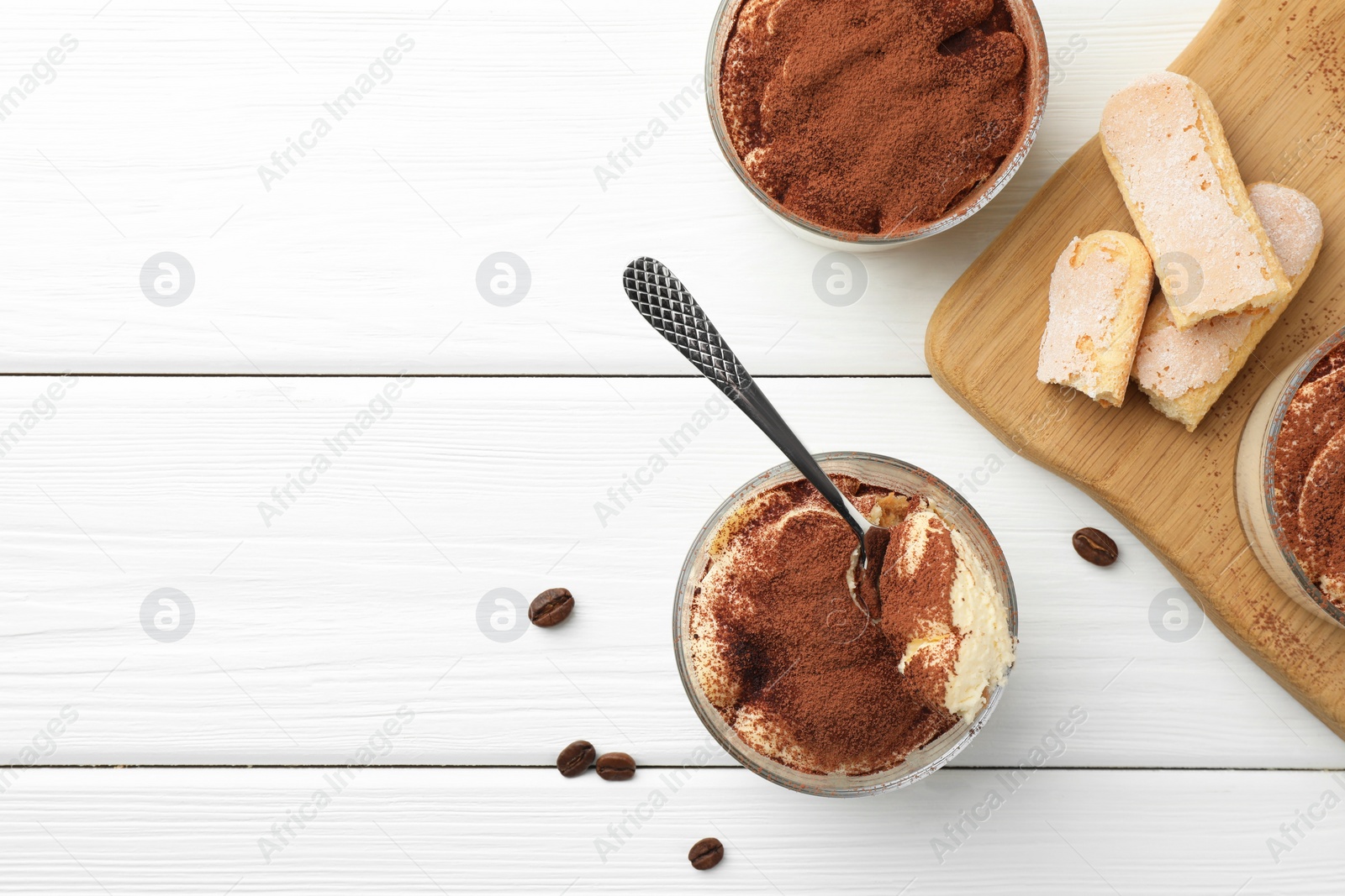 Photo of Delicious tiramisu in glasses, spoon, coffee beans and cookies on white wooden table, top view. Space for text